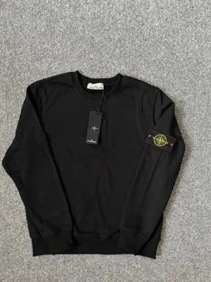 Svart sweatshirt från Stone Island - Säljer en svart sweatshirt från Stone Island med klassisk rund halsringning och ribbade muddar. Tröjan har den ikoniska Stone Island-loggan på vänster ärm och är tillverkad i mjukt bomullsmaterial. Perfekt för en stilren och avslappnad look.