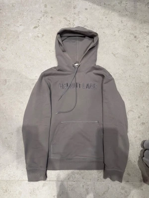 Grå hoodie från Helmut Lang - Snygg grå hoodie från Helmut Lang med broderad logga på bröstet. Klassisk känguruficka framtill, ribbade muddar och dragsko i huvan. Tillverkad i mjuk bomull för skön känsla och avslappnad passform.