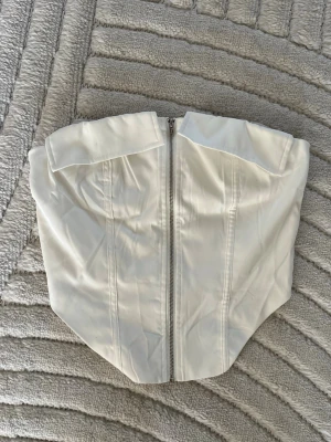 Vit korsett med dragkedja framtill - Säljer en stilren vit korsett med dragkedja framtill. Korsetten har en axelbandslös design och markerade sömmar som ger en snygg siluett. Materialet känns slätt och har en lätt glans. Perfekt att styla till olika looks. Aldrig använd!!