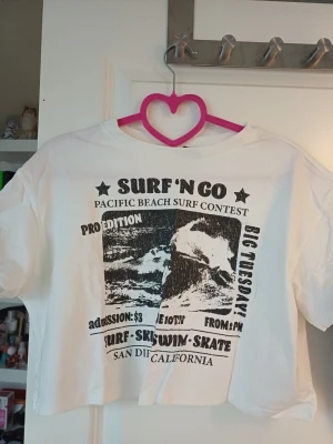 Vit croppad t-shirt med surftryck - Säljer en vit croppad t-shirt med svart surf-inspirerat tryck på framsidan. T-shirten har korta ärmar, rund halsringning och en avslappnad passform.