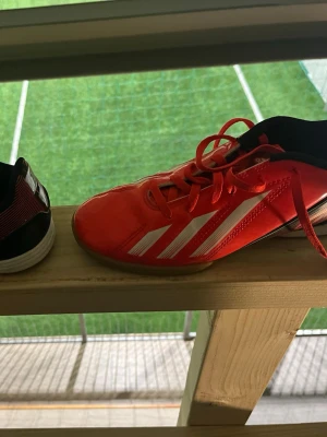 Röda Adidas fotbollsskor med vita ränder - Säljer ett par röda Adidas fotbollsskor med klassiska vita ränder på sidan. Skorna har snörning, svart insida och detaljer i svart och vitt på hälen. Yttermaterialet är syntetiskt och designen är sportig och modern, perfekt för dig som vill sticka ut på planen.