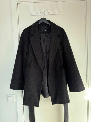 Svart kappa med bälte från Lager 157 - Stilren svart kappa från Lager157 med klassisk krage och avtagbart bälte i midjan. Kappan har har används ett par få gånger rak passform, långa ärmar och är helfodrad för extra komfort. Perfekt för kyliga dagar och enkel att matcha med olika outfits.