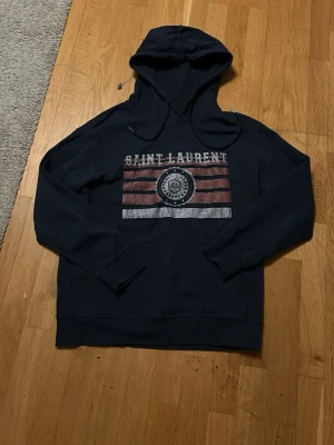 Mörkblå Saint Laurent hoodie - Snygg mörkblå hoodie från Saint Laurent med tryck framtill i vitt och rött. Klassisk huva med dragsko och magficka. Perfekt för dig som vill ha en stilren och trendig look med streetwear-vibe.