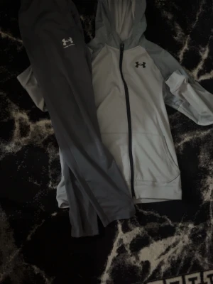 Under Armour grå träningsset - Snyggt träningsset från Under Armour med grå zip-hoodie och matchande träningsbyxor. Hoodien har dragkedja, fickor och logga på bröstet. Byxorna är mörkgrå med resår i midjan och logga på låret. Perfekt för gymmet eller löprundan.