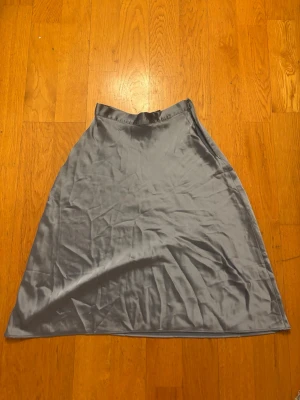 Grå satinlångkjol i A-linje - Snygg grå långkjol i satin med A-linjeformad siluett. Kjolen har en slät midja och ett elegant, glansigt material som ger en lyxig känsla. Perfekt för dig som vill ha en stilren och enkel look.