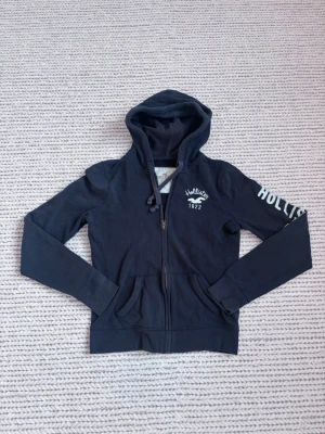 Mörkblå hoodie från Hollister - Mörkblå hoodie från Hollister med dragkedja framtill, broderad logga och texten 'Hollister 1922' på bröstet samt tryck på ena ärmen. Fint skick. Sitter som xs!