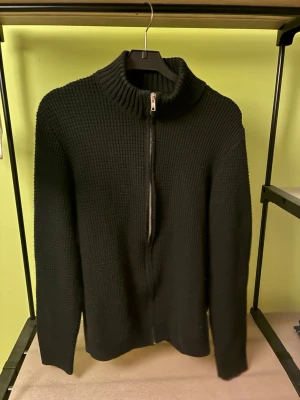 Svart stickad tröja med dragkedja - Säljer en svart stickad tröja från DeFacto med hög krage och dragkedja framtill. Tröjan har en grovstickad struktur och långa ärmar, perfekt för kyliga dagar. Enkel och stilren design som passar till det mesta.