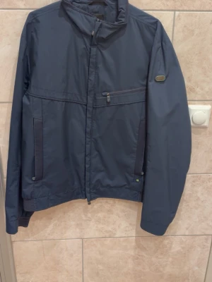 Mörkblå vindjacka med ståkrage - Snygg mörkblå vindjacka med ståkrage och dragkedja framtill. Jackan har en bröstficka med dragkedja, två sidofickor och ribbade muddar vid ärmslut och nederkant. Perfekt för blåsiga dagar och enkel att matcha till olika outfits.