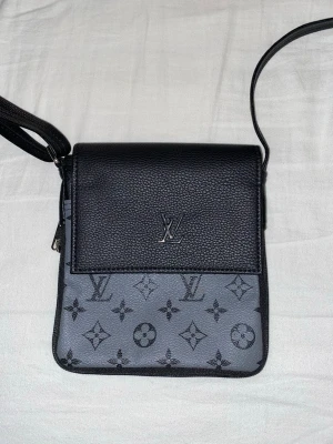 Louis Vuitton svart axelväska - Snygg axelväska från Louis Vuitton i svart och grått med klassiskt monogrammönster. Väskan har justerbar axelrem, lock med LV-logga i metall och är gjord i en kombination av slätt och präglat läder. Perfekt för dig som vill ha en stilren och trendig accessoar.