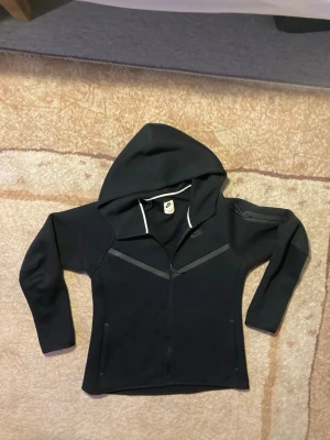 Svart hoodie från Nike med dragkedja - Säljer en svart hoodie från Nike med hel dragkedja framtill och huva. Hoodien har diskreta grå detaljer och Nike-logga på bröstet. Tillverkad i mjuk bomullsblandning och har långa ärmar samt fickor på sidorna. Perfekt för en sportig och avslappnad stil.