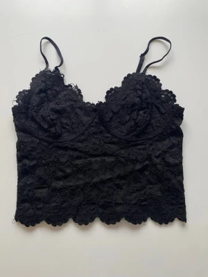 Svart spetslinnetopp med smala band - Snygg svart linnetopp med tunna axelband och vacker spets över hela toppen. Croppad modell med scallop-kant nertill och lätt vadderade kupor. Perfekt att styla till höga jeans eller kjol för en trendig look.