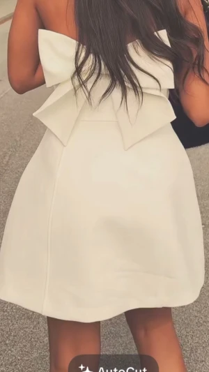 Vit axelbandslös kortklänning med rosett - Supersöt vit axelbandslös kortklänning med stor dekorativ rosett i ryggen. Klänningen har en lätt utsvängd kjol och en stilren, elegant look som passar perfekt till studenten och sommaren. Materialet känns slätt och har en lätt struktur.