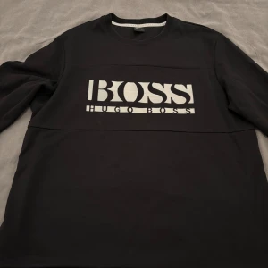 Svart Hugo Boss tröja - Svart sweatshirt från Hugo Boss.  Klassisk modell med rund halsringning och vit, broderad logotyp på bröstet. Ribbade muddar vid ärmslut och nederkant samt diskret QR-koddetalj i sidan. Mjuk och bekväm kvalitet.  Endast använd ett fåtal gånger och i mycket bra skick.