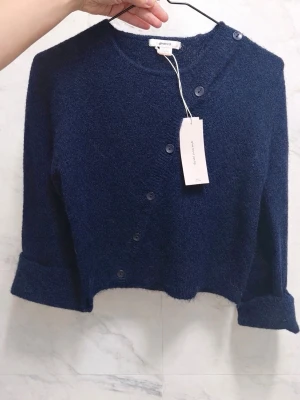 Mörkblå kofta från Gina tricot - Trendig mörkblå cropped kofta från Gina tricotmed rund halsringning och knappar framtill. Koftan har långa ärmar med breda uppvik och är tillverkad i ett mjukt, fluffigt material som ger en avslappnad look. Perfekt att styla över en topp eller klänning.