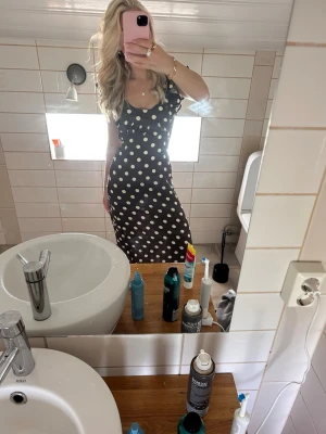 Svart prickig långklänning - Snygg svart långklänning med vita prickar och v-ringad hals. Klänningen har smala axelband och figurnära passform som framhäver siluetten. Perfekt för dig som vill sticka ut med ett klassiskt mönster.