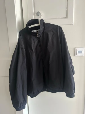 Svart jacka Acne Studios 1996 - Säljer en svart bomberjacka från Acne Studios med broderad text på ryggen: 'Acne Studios STOCKHOLM 1996'. Jackan har lång ärm, hög krage och dragkedja framtill. Perfekt oversized look och ribbade muddar vid ärmslut och nederkant.