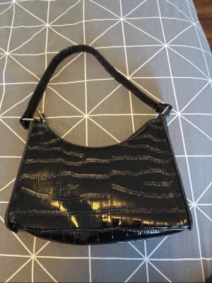 Svart axelväska i krokopräglat material - Snygg svart handväska med croco-mönstrad yta och kort axelrem. Väskan har en stilren, halvmåneformad design och stängs med dragkedja. Perfekt accessoar för att lyfta vilken outfit som helst.