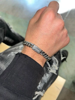 Kedjearmband i silver från BOSS (går att förhandla) - Snyggt kedjearmband i silverfärgad metall från BOSS. Armbandet har en stilren, rektangulär bricka med BOSS-loggan ingraverad. Länkarna är breda och ger ett modernt och trendigt intryck. Perfekt accessoar för att lyfta din outfit.