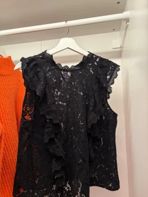 Svart spetsblus med volangdetaljer - Säljer en svart blus i spets med volangdetaljer längs axlar och framstycke. Blusen är ärmlös och har en rund halsringning med dekorativ spetskant. Perfekt för dig som gillar en romantisk och edgy stil.