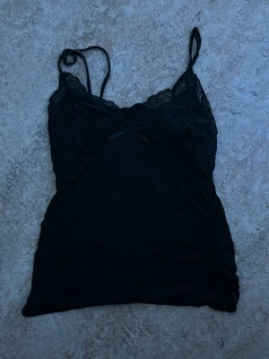 Svart spetslinnetopp med rosett - Snygg svart linnetopp med tunna axelband och spetsdetaljer upptill. Toppen har en liten rosett framtill och är figurnära i modellen. Perfekt att styla lager på lager eller bära som den är.