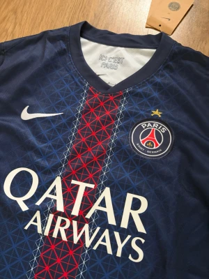PSG Vitinha Fotbollströja - Snygg mörkblå PSG matchtröja 25/26 med röd och vit detalj framtill, officiellt klubbmärke och Qatar Airways-tryck. På ryggen står det Vitinha och nummer 17. Tillverkad i lätt och ventilerande material, perfekt för fotboll eller träning. På axeln finns Champions League loggan. Tröjan är storlek S och är helt ny (lappen fortfarande på).                Skriv om ni har några funderingar!😄