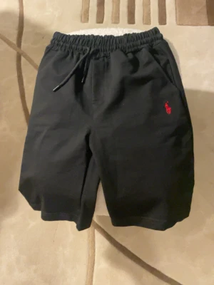 Svarta shorts från Polo Ralph Lauren - Svarta shorts från Polo Ralph Lauren med elastisk midja och snörning. Klassisk röd logga broderad på vänster sida. Tillverkade i mjuk bomull, perfekta för en avslappnad stil. Två sidofickor och normal passform.