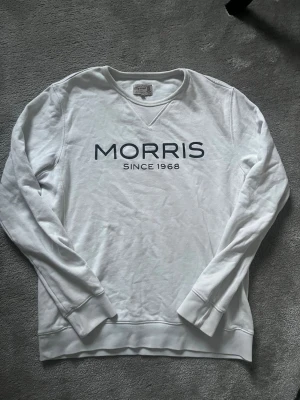 Vit långärmad tröja från Morris - Vit långärmad tröja från Morris med tryckt logga 'MORRIS SINCE 1968' på bröstet. Klassisk rund halsringning och ribbade muddar vid ärmslut och nederkant. Perfekt för en clean och stilren look.
