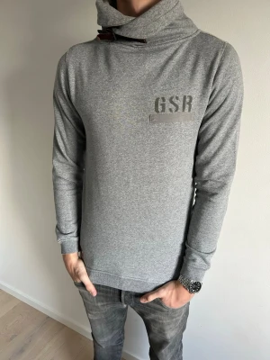 Grå hoodie med hög krage från GSR - Snygg grå hoodie från G-Star Raw med hög krage och schyssta spännen vid halsen. Tröjan har tryckt logga på bröstet och är tillverkad i mjukt, stretchigt material. Långa ärmar och ribbade muddar ger en clean look. Perfekt för dig som vill ha en snygg hoodie som passar till allt:) Tröjan är storlek S men känns absolut mer som en M. Hör mer än gärna av er vid frågor😁