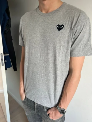 Grå Comme des Garçons T-shirt - Snygg grå T-shirt från Comme des Garçons med det ikoniska svarta hjärta-ögon-loggan på bröstet. Perfekt till jeans eller shorts för en clean och stilren look. Passa på att köpa en snygg t shirt nu till sommarn☺️ Hör av er om ni undrar något!