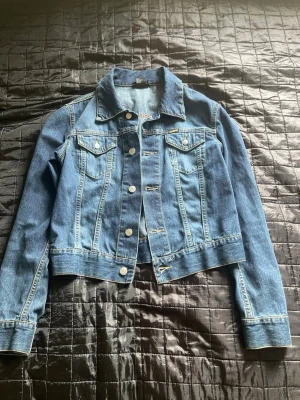 Blå klassisk jeansjacka från Diesel - Snygg blå jeansjacka med silverfärgade knappar och två bröstfickor med lock. Jackan har en klassisk krage och markerade sömmar som ger en tidlös look. Perfekt att slänga över en hoodie eller t-shirt för en chill stil. Storleken är M i kvinnstorlek 