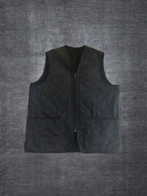 Svart quiltad dunväst med dragkedja från Barbour - Stilren svart dunväst med quiltat mönster och dragkedja framtill från Barbour. Västen har två stora fickor och en klassisk rak passform. Perfekt att slänga över en hoodie eller tröja för extra värme. Materialet är glansigt och lätt, vilket gör den enkel att bära.