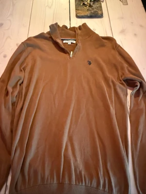 Brun tröja med dragkedja från U.S. Polo Assn. - Snygg brun tröja från U.S. Polo Assn. med halv dragkedja och ribbad krage. Tröjan har lång ärm och en broderad logga på bröstet. Perfekt till jeans eller chinos för en avslappnad men stilren look.