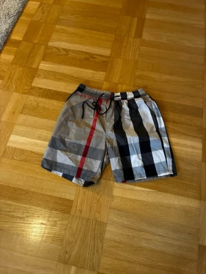 Rutig badshorts med snörning - Har bara används som shorts!!!Säljer ett par klassiska badshorts från Burberry med rutigt mönster i svart, grått, vitt och en röd rand. Shortsen har snörning i midjan och är tillverkade i ett lätt syntetmaterial som torkar snabbt. Perfekta för sommaren!