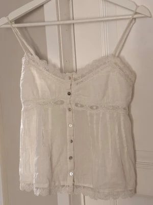 Vit spetslinnetopp från Zara - Säljer en söt vit linnetopp med tunna axelband och dekorativa spetsdetaljer längs halsringning och nederkant. Säljs inte längre. Skriv vid frågor💕