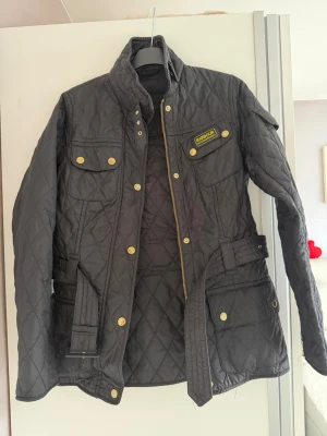 Svart quiltad vindjacka från Barbour - Stilren svart quiltad vindjacka från Barbour med bälte i midjan, guldiga knappar och flera fickor framtill. Klassisk design med hög krage och Barbour-logga på bröstet. 