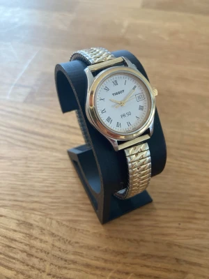 1990 Tissot Safirglas Date 27mm Damur - Tissot PR 50 damklocka med kvartsrörelse. Klockan har en vit urtavla med romerska siffror och en datumvisning. Boetten är i stål med guldtonade detaljer runt urtavlan och på kronan. Armbandet är ett flexibelt länkarmband i stål och guldtonat. Baksidan av boetten är märkt med "WATERRESISTANT 50M", "SWISS PACK", "SAPPHIRE CRYSTAL" och "JT32/232K". Nära nyskick enligt bilder, inga repor, behöver bara nytt batteri.   Begagnat värde: < 1500 SEK Mitt pris: 689 SEK  Skickas alltid snabbt och spårbart. 