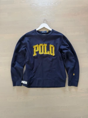 Mörkblå sweatshirt från Polo Ralph Lauren - Cool mörkblå sweatshirt med stort gult POLO-tryck på bröstet och liten broderad logga på ärmen. Klassisk rund halsringning och ribbade muddar. Perfekt för dig som gillar sportig och avslappnad stil.