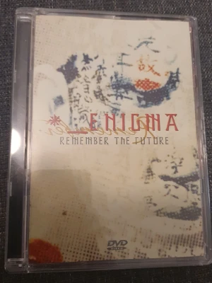 Enigma Remember The Future DVD - Enigma Remember The Future DVD med abstrakt konst på omslaget och text i rött och svart. DVD-fodralet är genomskinligt och innehåller information om låtar och produktion på baksidan. Perfekt för dig som gillar musikdokumentärer och samlar på DVD.