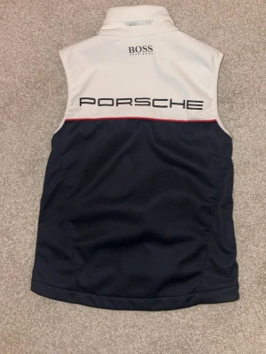 Porsche x BOSS väst i marinblått och vitt - Snygg väst från BOSS i samarbete med Porsche. Marinblå nedtill och vit upptill med röd detaljlinje. Dragkedja framtill, hög krage och loggor på både fram- och baksida. Perfekt för dig som gillar sportig och stilren design. Storlek XS kan passa S