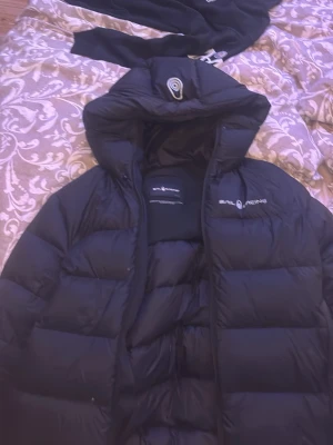 Svart dunjacka från Sail Racing - Säljer en svart dunjacka från Sail Racing med huva och diskret logga på bröstet. Jackan har en puffig design med breda kanaler och dragkedja framtill. Perfekt för kalla vinterdagar och riktigt snygg till streetwear-stilen.