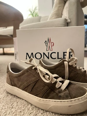 Bruna Moncler sneakers i mocka - Säljer ett par snygga bruna Moncler sneakers i mocka med vita sulor och matchande beige skosnören. Klassisk låg modell med diskreta sömdetaljer och stilren design. Perfekta för dig som vill ha en clean och exklusiv look.