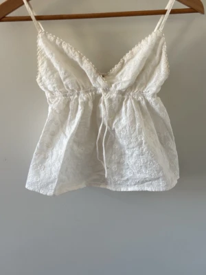 Vit broderad linnetopp med spets - Söt vit linnetopp med tunna axelband, broderade blommor och spetsdetaljer. Toppen har v-ringning, justerbara axelband och en liten knytning framtill. Från subdued 