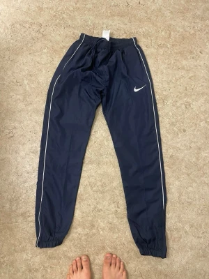 Marinblå träningsbyxor från Nike - Snygga marinblå träningsbyxor från Nike med vita detaljer längs benen och klassisk Nike-logga på låret. Byxorna har resår i midjan och vid bensluten för skön passform. Perfekta för träning eller chill. Tillverkade i lätt och smidigt material.