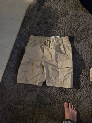 Beige cargoshorts med snörning - Säljer ett par beige cargoshorts med snörning i midjan och stora sidofickor. Perfekta för sommaren och riktigt sköna med avslappnad passform. Passar dig som gillar praktiska detaljer och enkel stil.