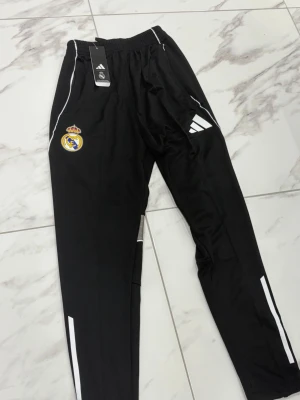 Svarta Real Madrid träningsbyxor Adidas - Snygga svarta träningsbyxor från Adidas med Real Madrid-logga på ena benet och vita detaljer längs sidorna. Byxorna har resår i midjan, smal passform och är tillverkade i ett lätt, sportigt material. Perfekta för fotbollsträning eller gymmet.