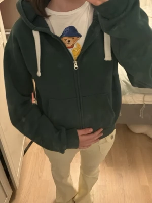 Mörkgrön zip hoodie med huva - Mysig mörkgrön hoodie med dragkedja framtill och stora fickor. Har vit dragsko i huvan och ribbade muddar vid ärmar och nederkant. Perfekt för en avslappnad och chill stil. Materialet känns mjukt och bekvämt, troligen bomullsblandning.