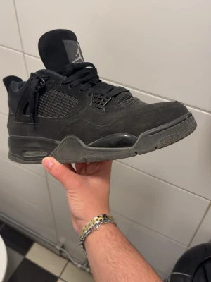 Svarta Air Jordan 4 - Snygga helsvarta Air Jordan 4 Retro sneakers med meshpaneler, mockadetaljer och klassisk snörning. Skon har en chunky sula och ikoniska detaljer som plastvingar och Jumpman-logga på plösen. Perfekt för dig som gillar streetwear och vill ha en stilren look.