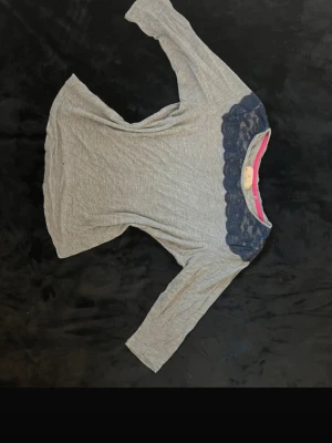 Grå topp med spetsdetalj - Snygg grå topp med mörkblå spets över axlarna och trekvartsärm. Mjuk och skön i bomullsmix, perfekt till jeans eller kjol. Spetsen ger en extra touch och toppen har en normal passform. Finns ett litet hål men inget som syns. 