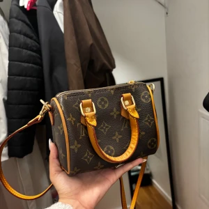 Mini Handväska - Säljer en brun minihandväska från med klassiskt monogrammönster och detaljer i ljusbrunt läder. Väskan har både handtag och avtagbar axelrem i samma färg, samt guldfärgade metalldetaljer. Perfekt för dig som gillar lyxiga accessoarer.