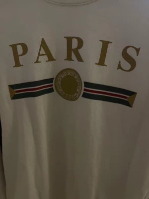 Vit sweatshirt med PARIS-tryck  - Säljer en vit sweatshirt med stort PARIS-tryck i guld på bröstet och dekorativt band i grönt, rött och vitt. Tröjan har rund halsringning och långa ärmar. Perfekt för dig som gillar streetstyle och vill ha en snygg statementtröja.